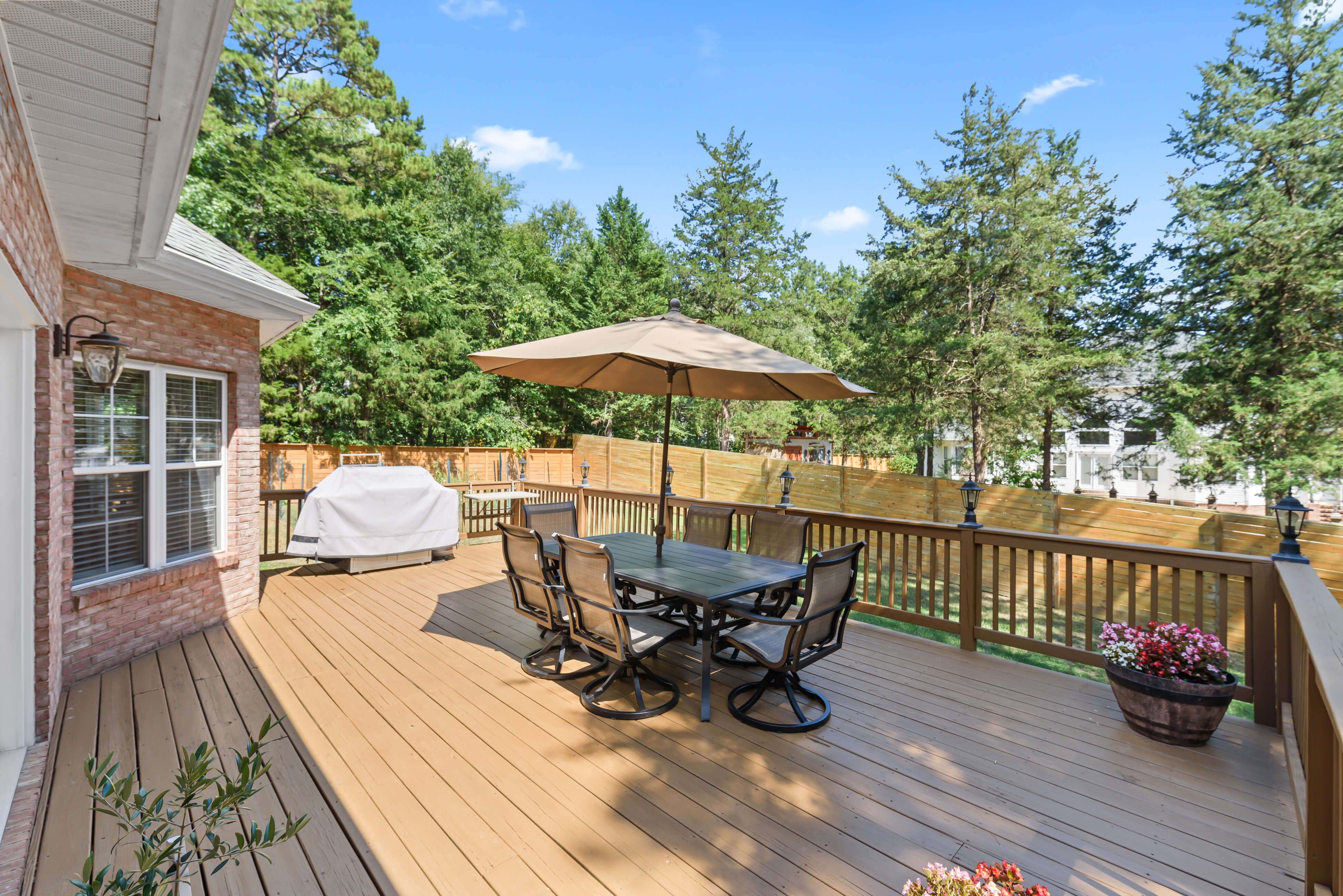Spacious backyard deck with dining table and umbrella, grill station, privacy fence, and wooded backdrop.