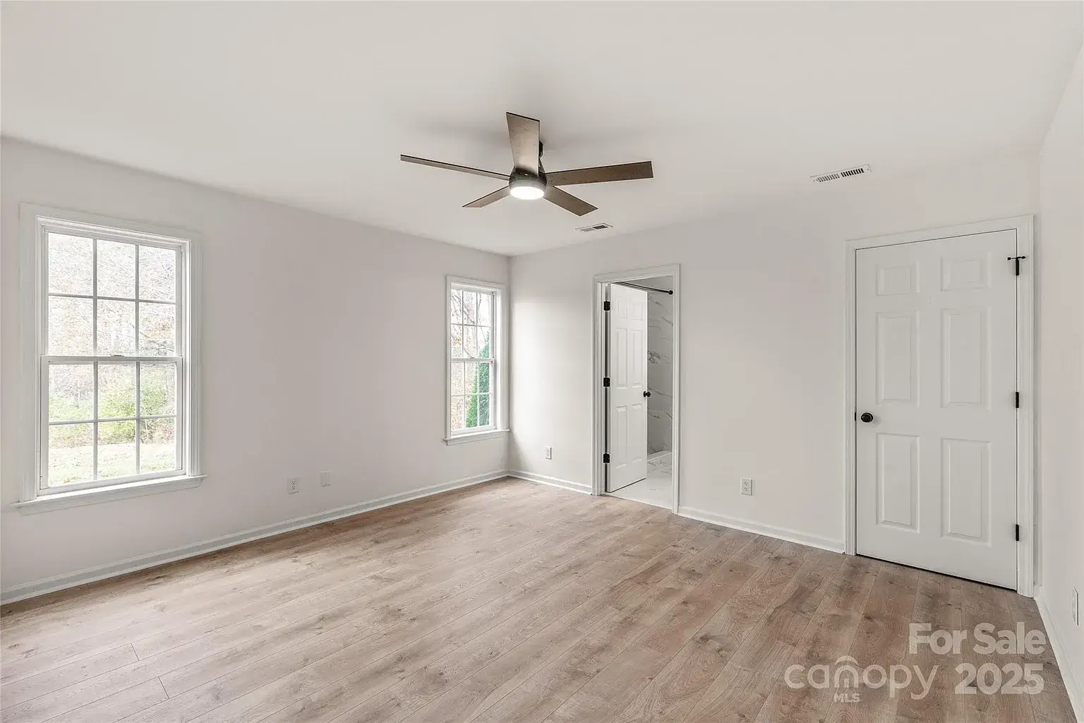 Primary bedroom with light wood flooring, ceiling fan, two windows, and open doorway to en-suite bathroom.