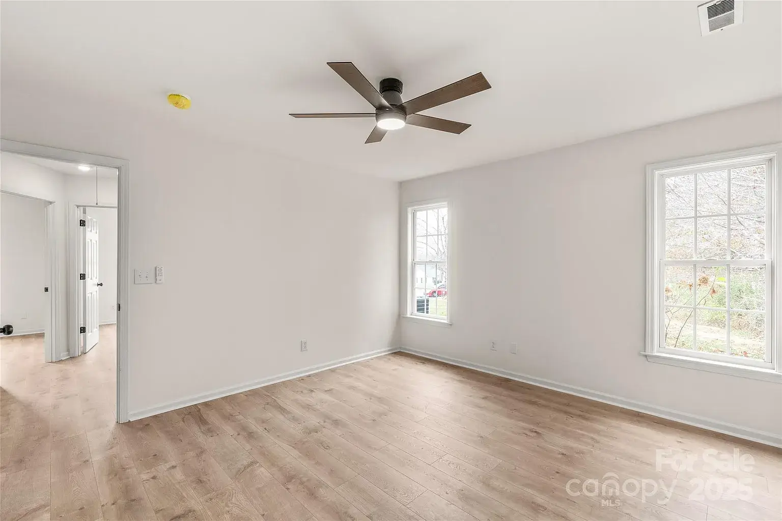 Bedroom with light wood flooring, ceiling fan, two windows, and doorway to hallway.