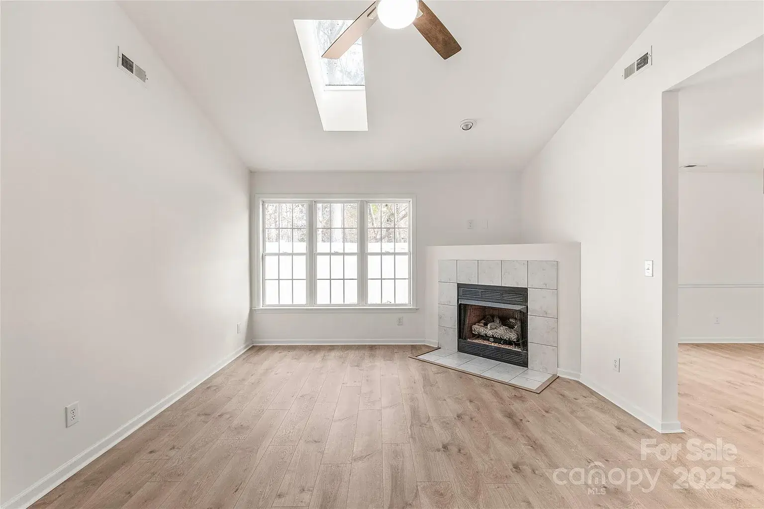 Living room with vaulted ceiling, skylight, ceiling fan, large window, gas fireplace, and wood flooring.