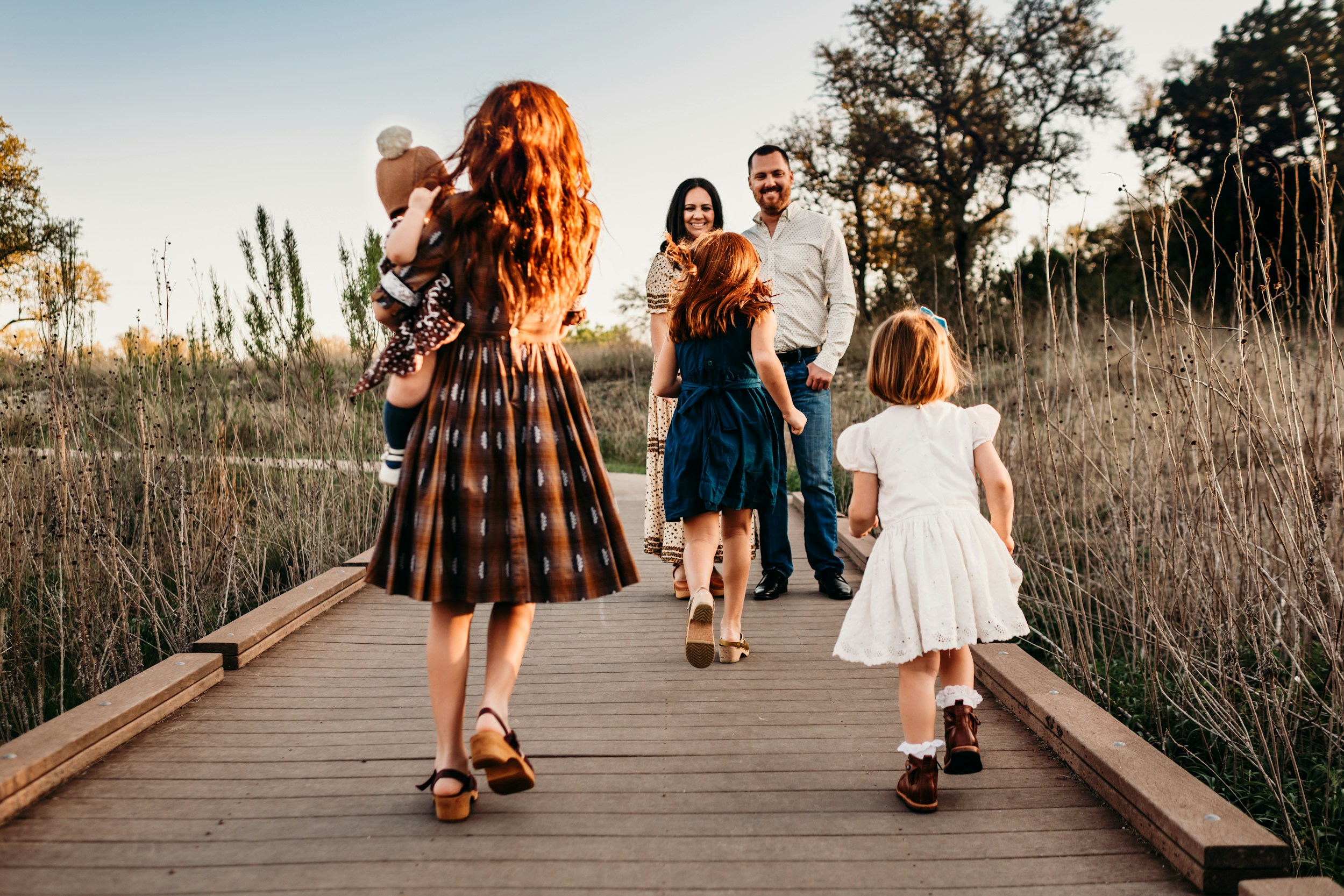 Family with young children walking on a wooden boardwalk through a field at sunset.