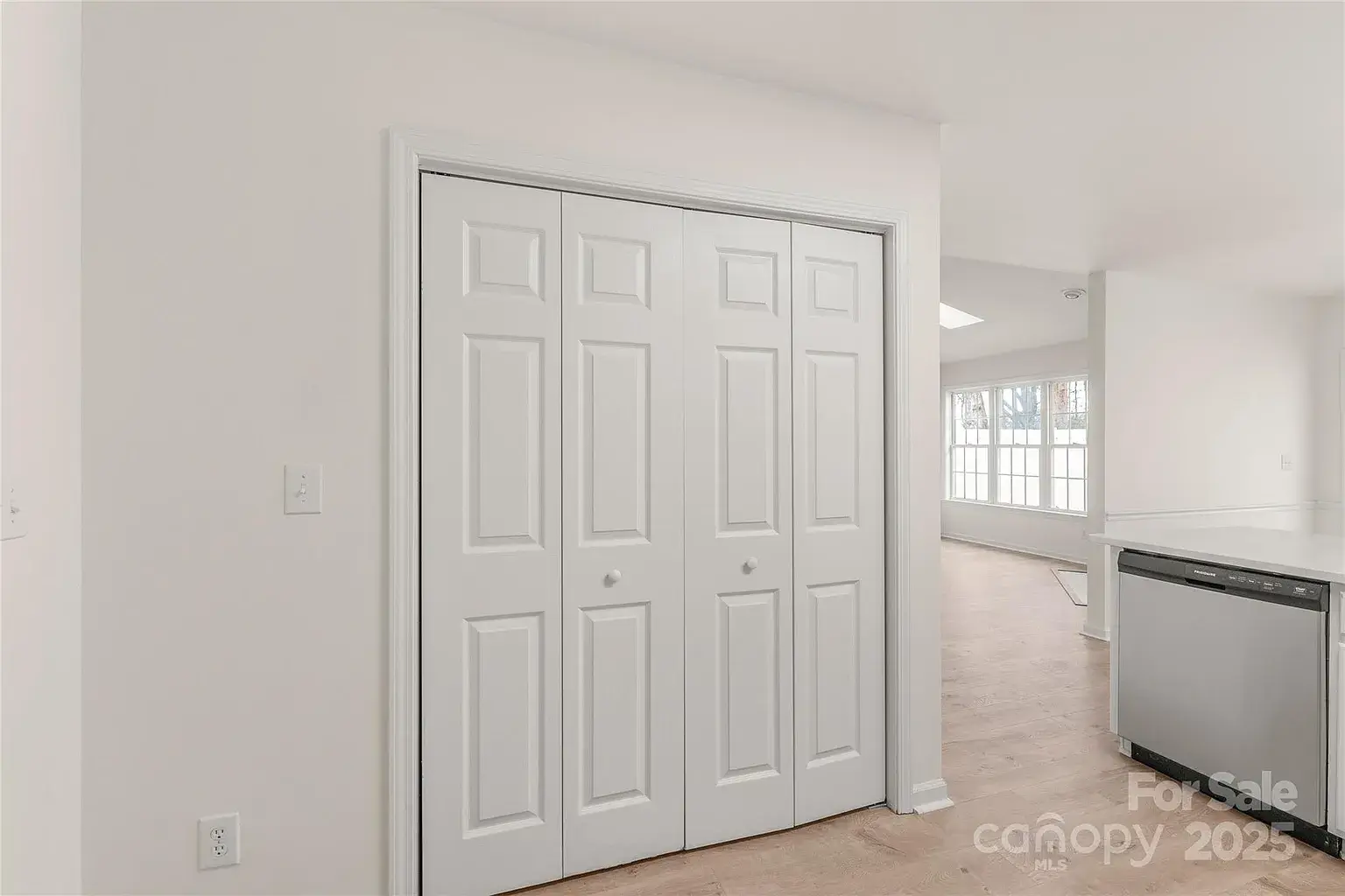 White double-door interior closet near kitchen and living area with light wood flooring.