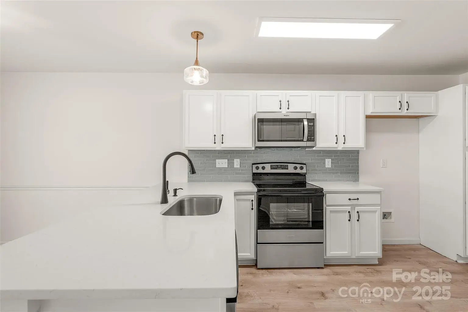 Bright white kitchen with quartz peninsula, undermount sink, stainless steel range and microwave, gray tile backsplash, and modern pendant lighting.