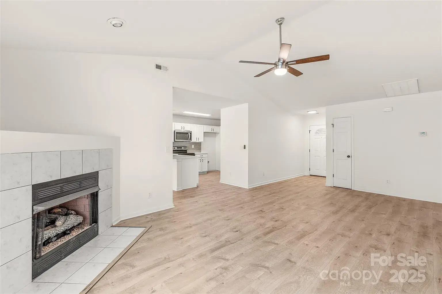 Open living room with gas fireplace, vaulted ceiling, ceiling fan, wood flooring, and view into white kitchen.