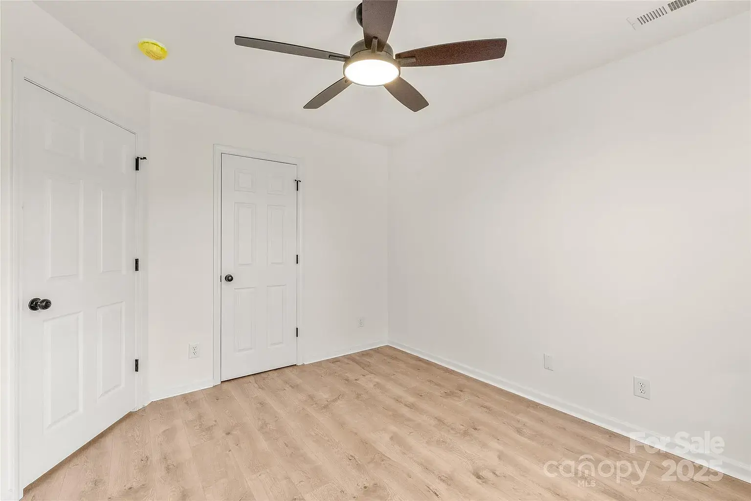 Empty bedroom with hardwood floors, white walls, ceiling fan with light, and double doors.