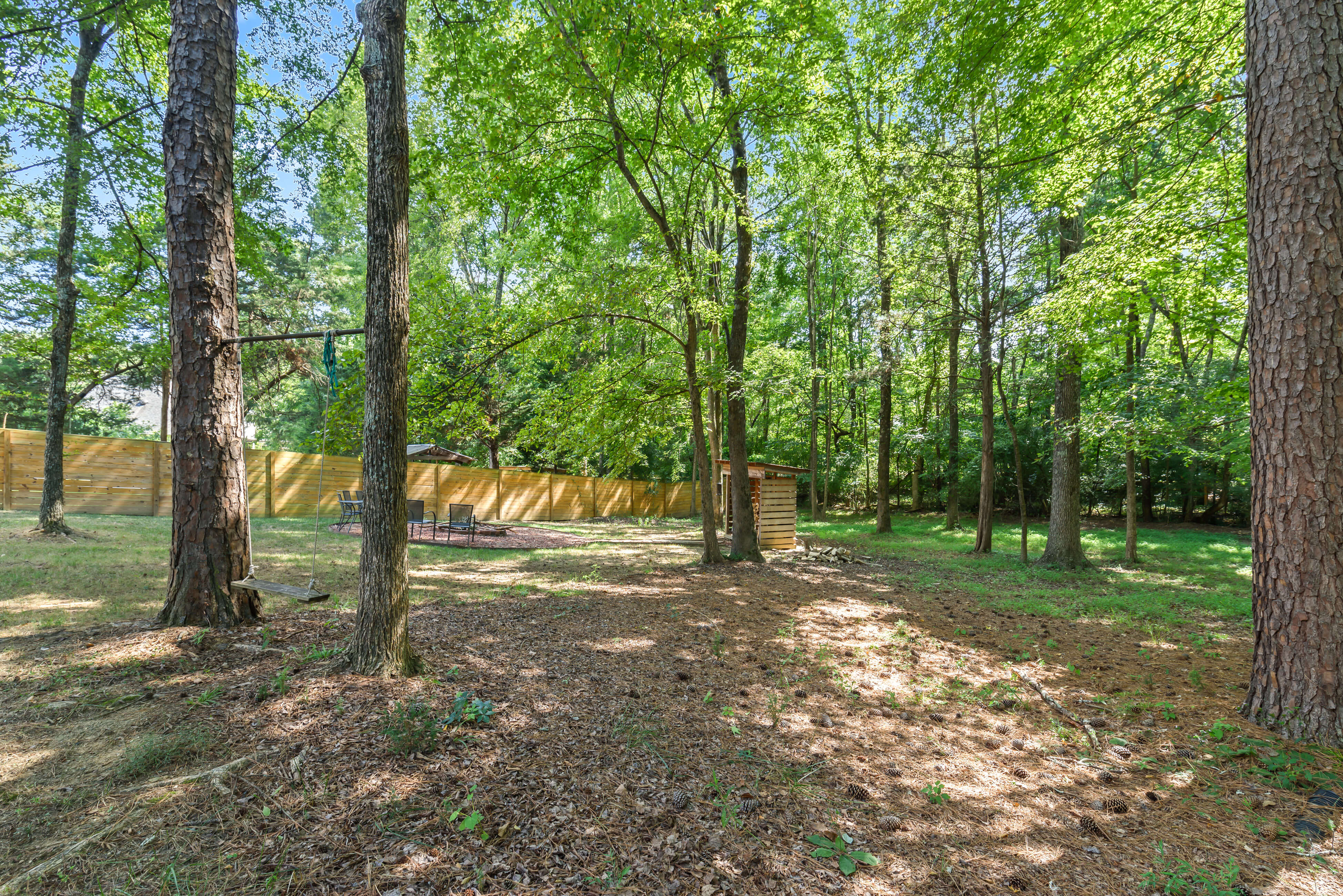 Wooded yard with rustic tree swing, privacy fence, and wood storage shed near the fire pit area.