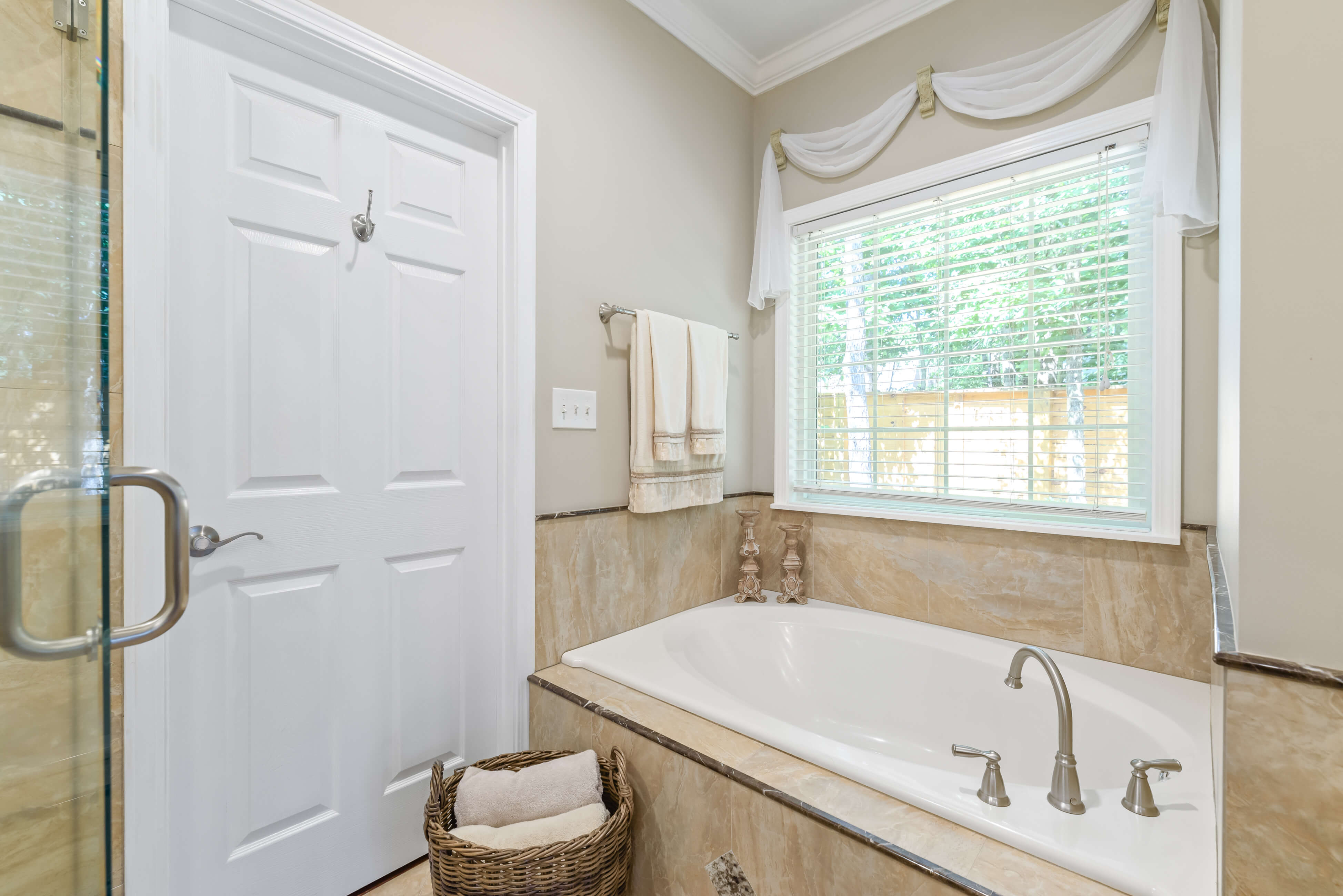 Soaking tub beneath window next to glass shower, with towel rack and basket