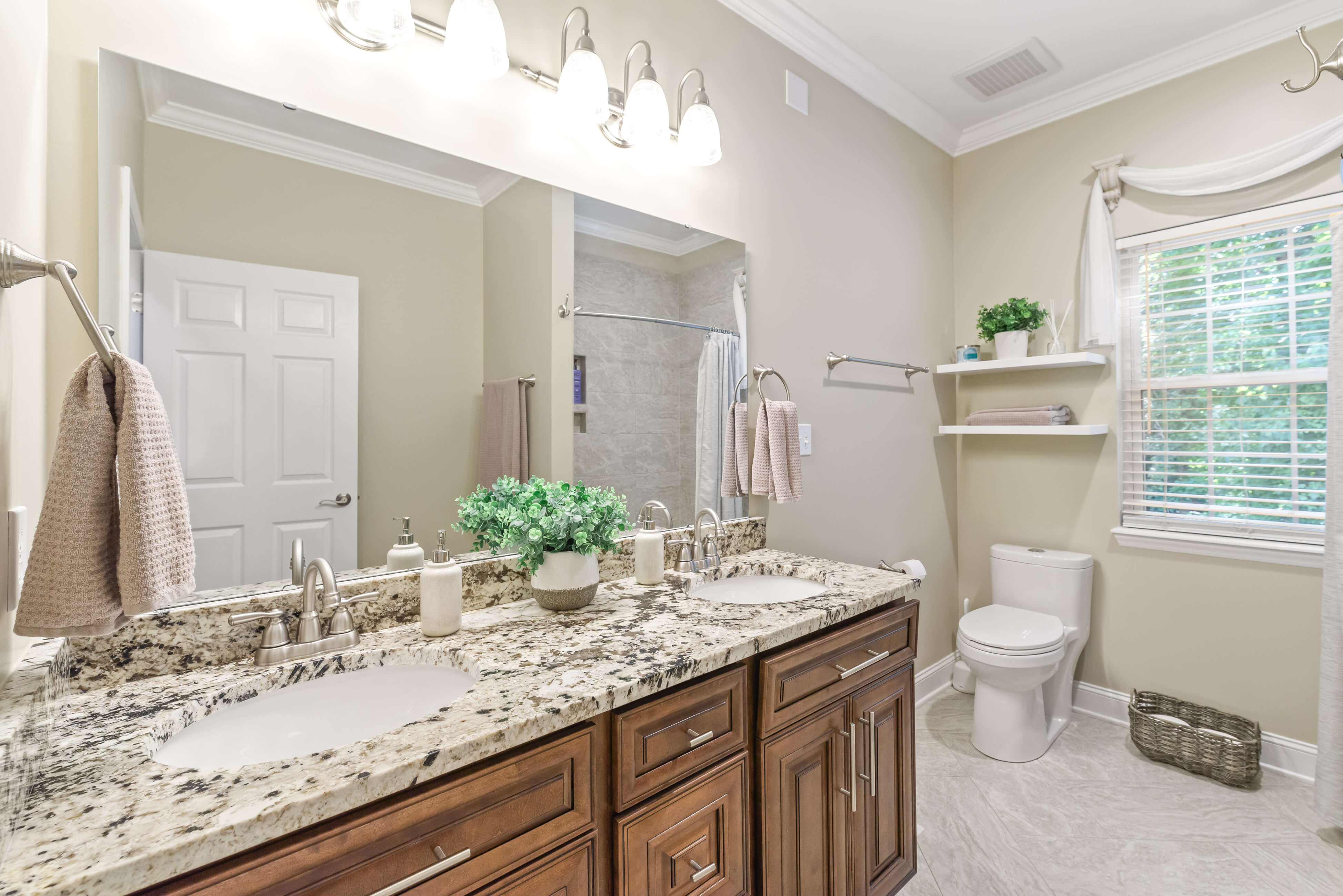 Bathroom with granite double vanity, large mirror, floating shelves, toilet, and window