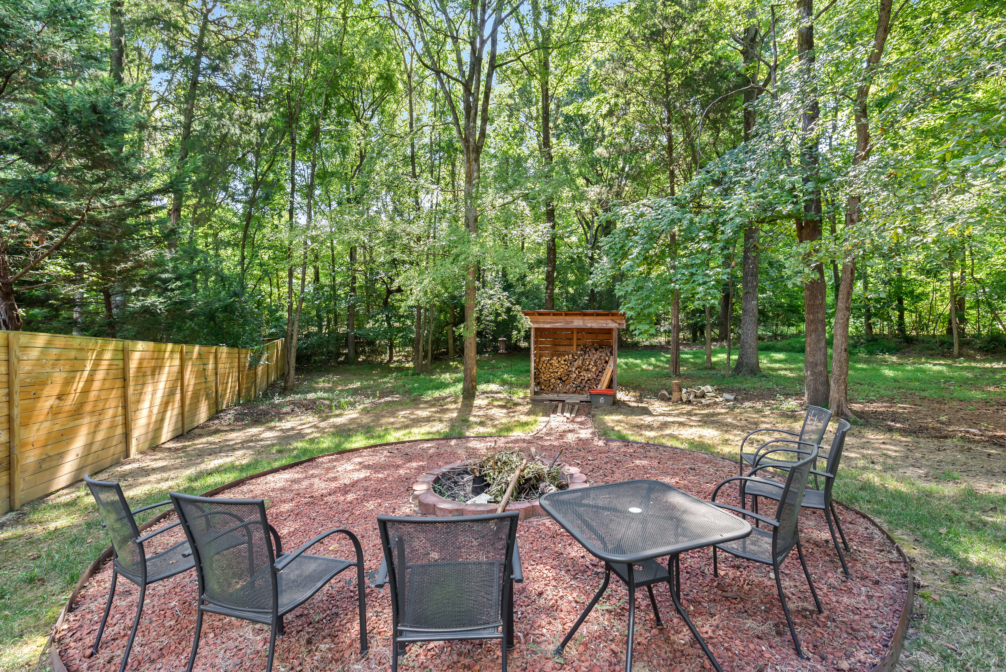 Backyard fire pit circle with chairs on red gravel, wood storage shed, and wooded fence line.