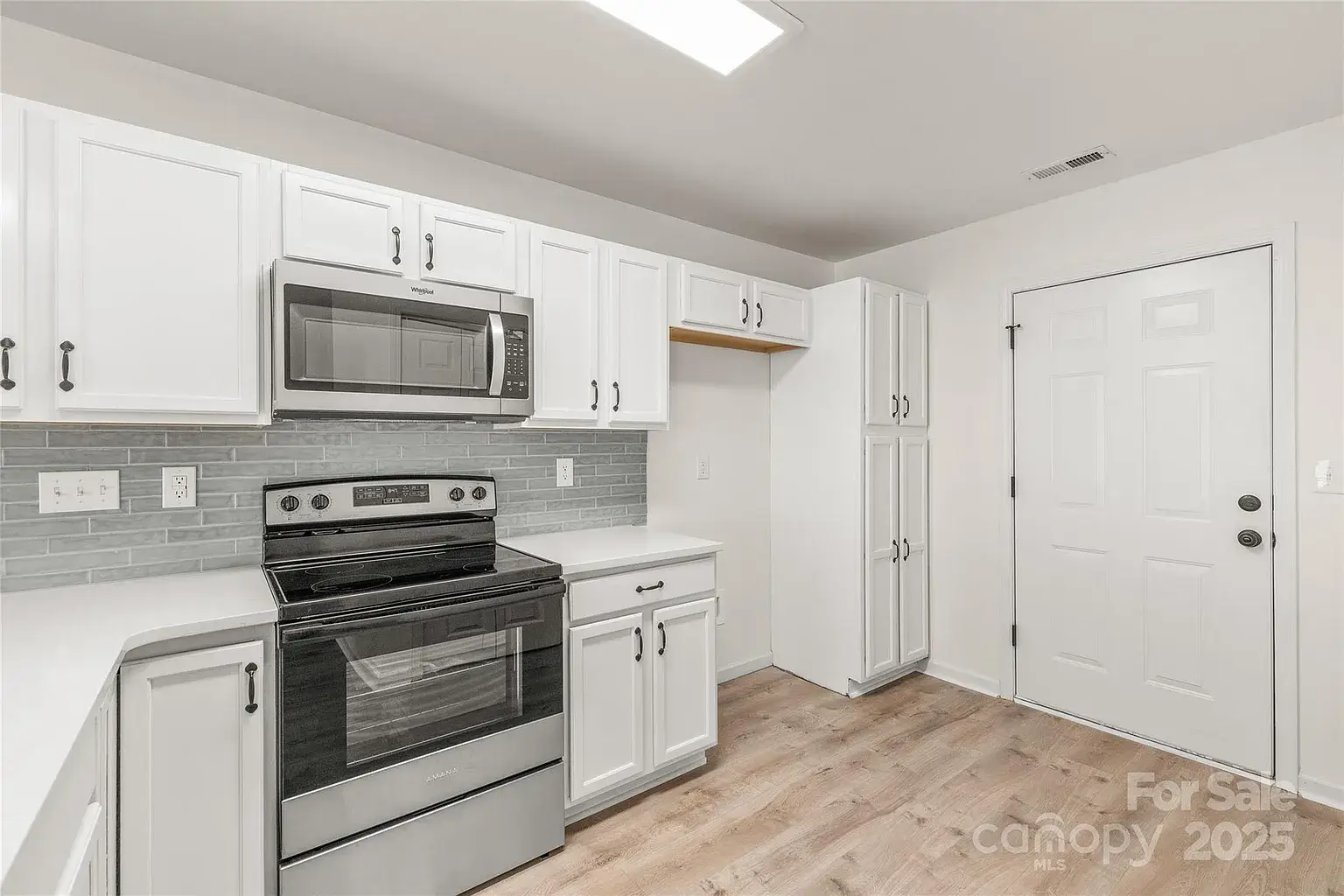 White kitchen with stainless steel range and microwave, gray subway tile backsplash, tall pantry cabinets, and adjacent exterior door.