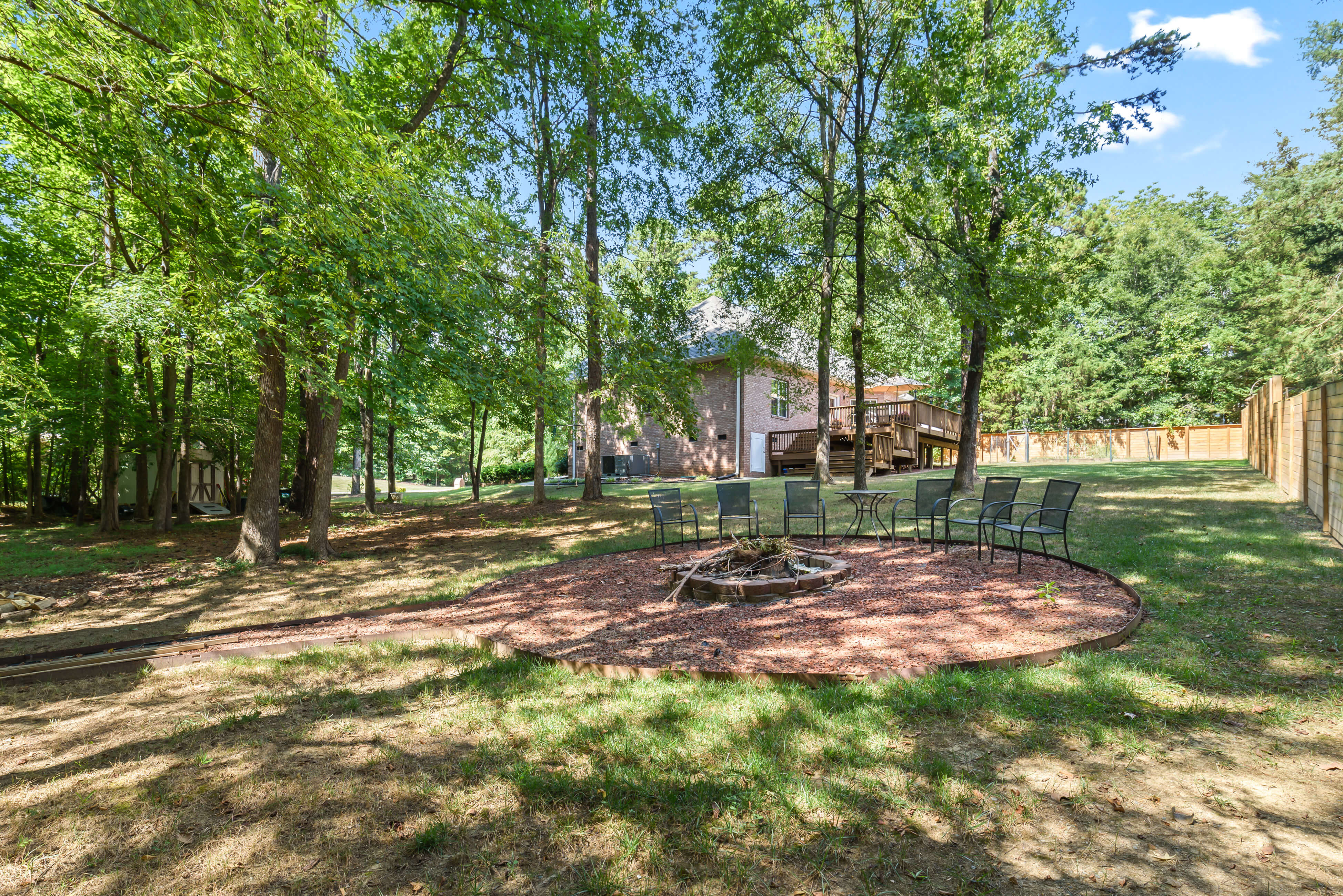 Wide view of wooded backyard with fire pit seating area and the home’s deck visible in the distance.
